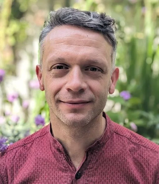 Simon Shaw, a white man with grey hair smiles gently at the camera wearing a red shirt